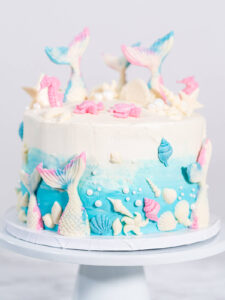 Meerjungfrauen-Torte auf weißem Tortenständer, oben weißes Frosting, unten blauer Farbverlauf, mit weißen Schokoladen-Flossen, rosa Seepferdchen, Muscheln und weißen Zuckerperlen