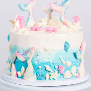 Meerjungfrauen-Torte auf weißem Tortenständer, oben weißes Frosting, unten blauer Farbverlauf, mit weißen Schokoladen-Flossen, rosa Seepferdchen, Muscheln und weißen Zuckerperlen
