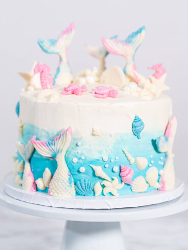 Meerjungfrauen-Torte auf weißem Tortenständer, oben weißes Frosting, unten blauer Farbverlauf, mit weißen Schokoladen-Flossen, rosa Seepferdchen, Muscheln und weißen Zuckerperlen