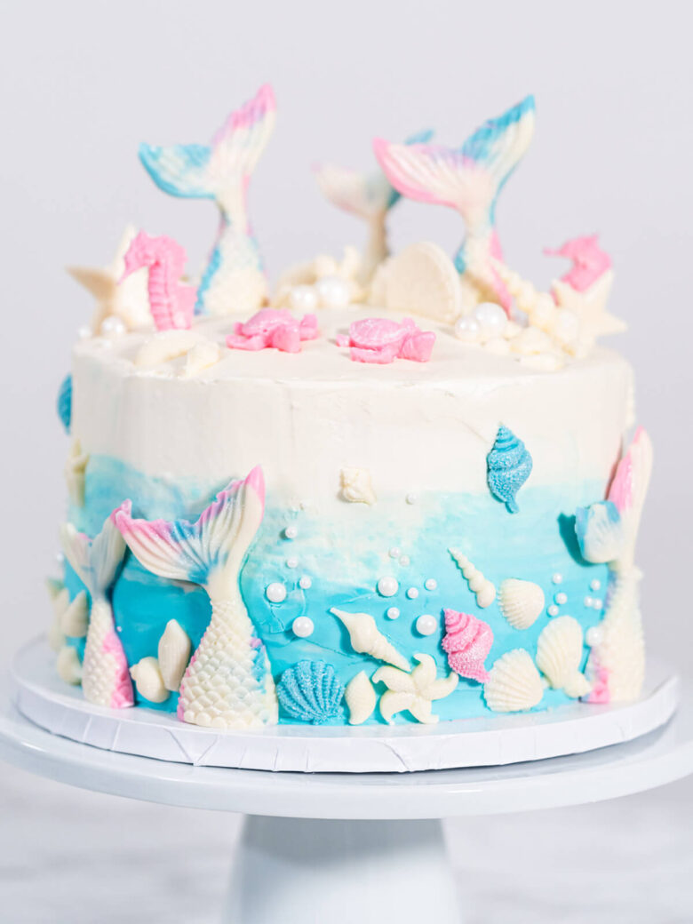 Meerjungfrauen-Torte auf weißem Tortenständer, oben weißes Frosting, unten blauer Farbverlauf, mit weißen Schokoladen-Flossen, rosa Seepferdchen, Muscheln und weißen Zuckerperlen