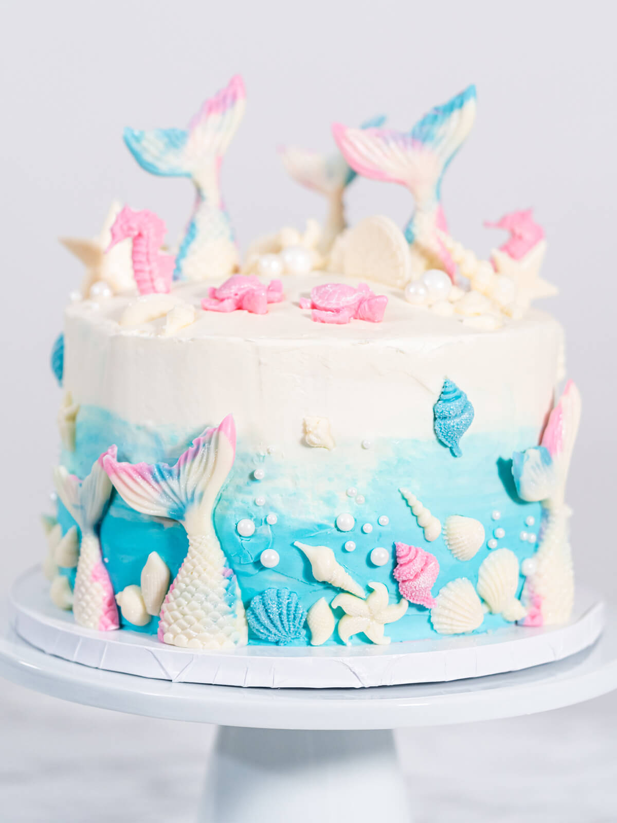 Meerjungfrauen-Torte auf weißem Tortenständer, oben weißes Frosting, unten blauer Farbverlauf, mit weißen Schokoladen-Flossen, rosa Seepferdchen, Muscheln und weißen Zuckerperlen