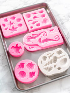 Verschiedene rosa und weiße Silikonformen für Muscheln, Seesterne, Seepferdchen und eine Schildkröte auf einem Backblech