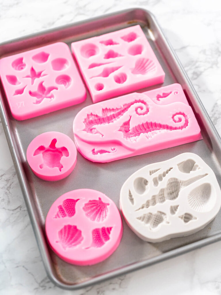 Verschiedene rosa und weiße Silikonformen für Muscheln, Seesterne, Seepferdchen und eine Schildkröte auf einem Backblech