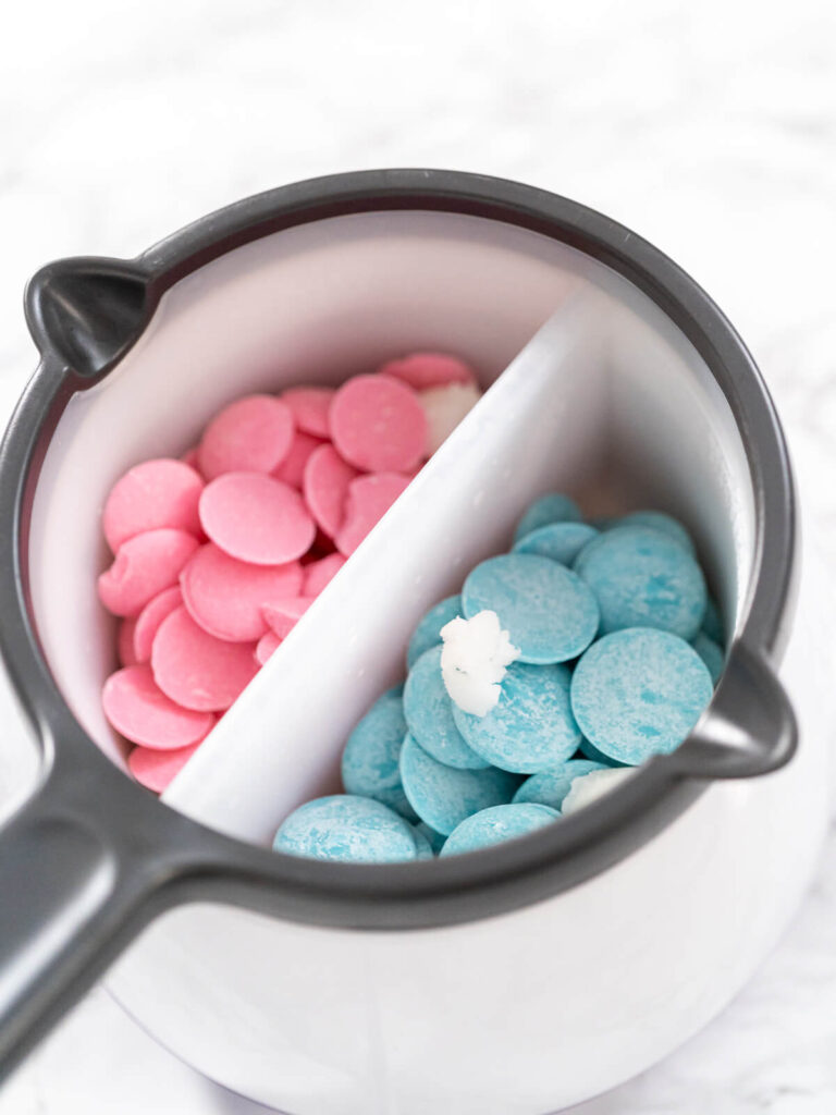 Schmelztopf mit rosa und blauen Schmelzglasur-Drops, getrennt durch einen Steg, dazwischen ein Stück Kokosöl