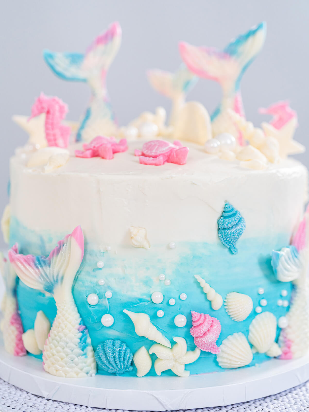 Meerjungfrauen-Torte schräg von oben auf weißem Tortenständer, drei aufrecht stehende Schokoladen-Flossen, rosa Seepferdchen und Schildkröten auf dem weißen Deckel