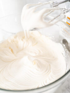 Weiße amerikanische Buttercreme in einer Glasschüssel, die Rührbesen des Handmixers ziehen weiche Spitzen aus der glänzenden Masse