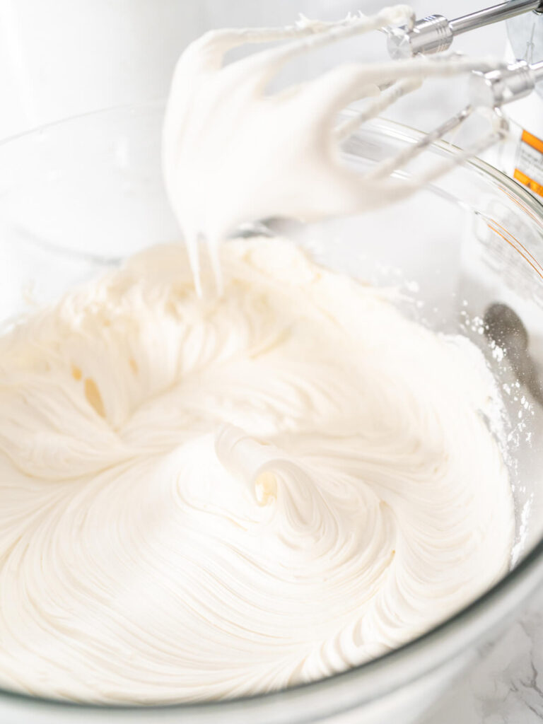 Weiße amerikanische Buttercreme in einer Glasschüssel, die Rührbesen des Handmixers ziehen weiche Spitzen aus der glänzenden Masse