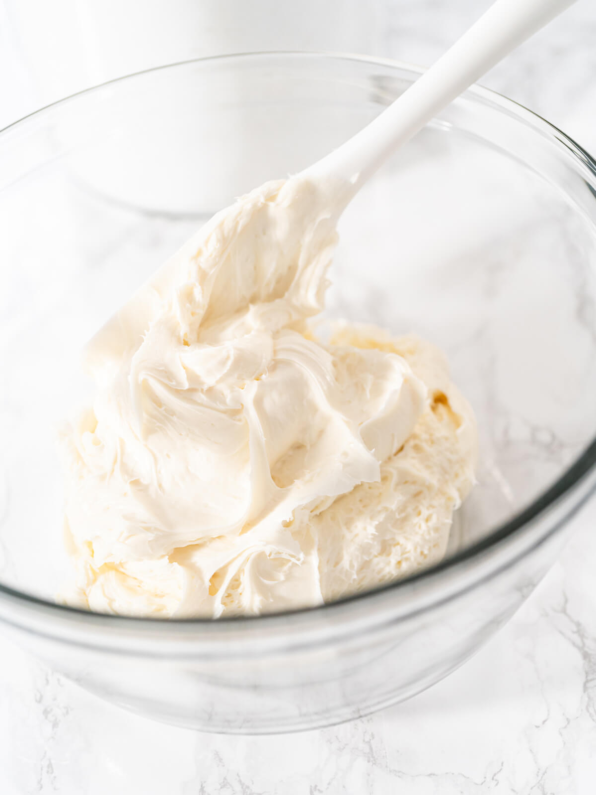 Cremige weiße Buttercreme in einer Glasschüssel, mit einem weißen Teigschaber glatt gerührt