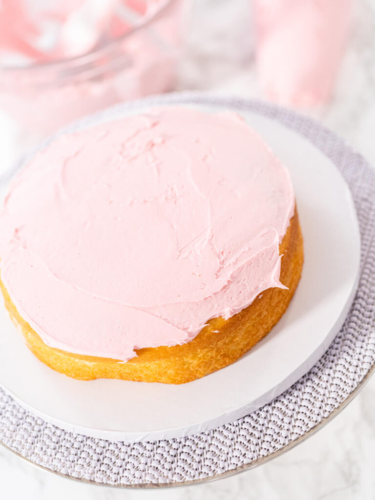 Erster goldbrauner Vanilleboden auf einer weißen Kuchenpappe, gleichmäßig mit rosa Buttercreme bestrichen