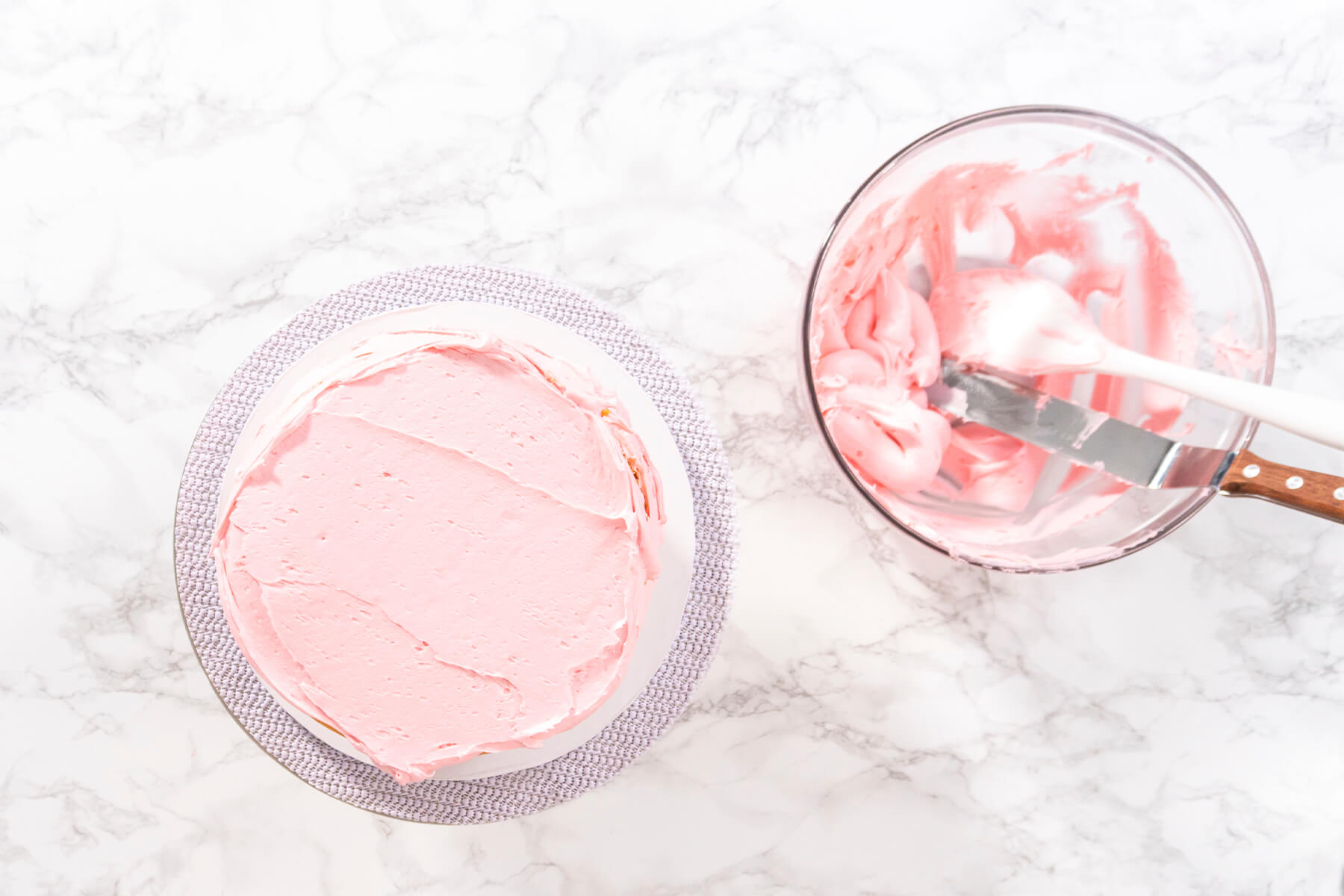Erster Tortenboden von oben, gleichmäßig mit rosa Buttercreme bestrichen, daneben eine Glasschüssel mit rosa Creme und einer Winkelpalette