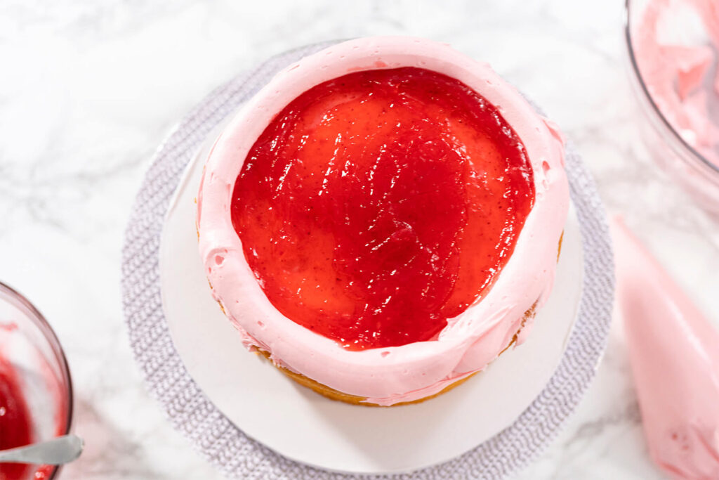 Tortenboden von oben mit einem geschlossenen Ring aus rosa Buttercreme am Rand, die Mitte mit glänzender roter Konfitüre gefüllt