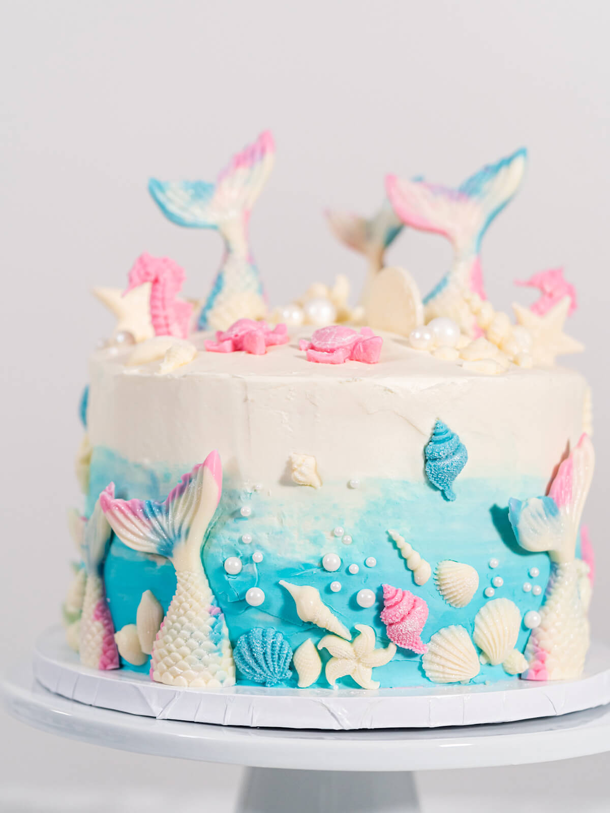 Vollständig dekorierte Meerjungfrauen-Torte von vorn, blauer Farbverlauf unten mit Muscheln, Seesternen und Zuckerperlen, oben weißer Deckel mit Flossen