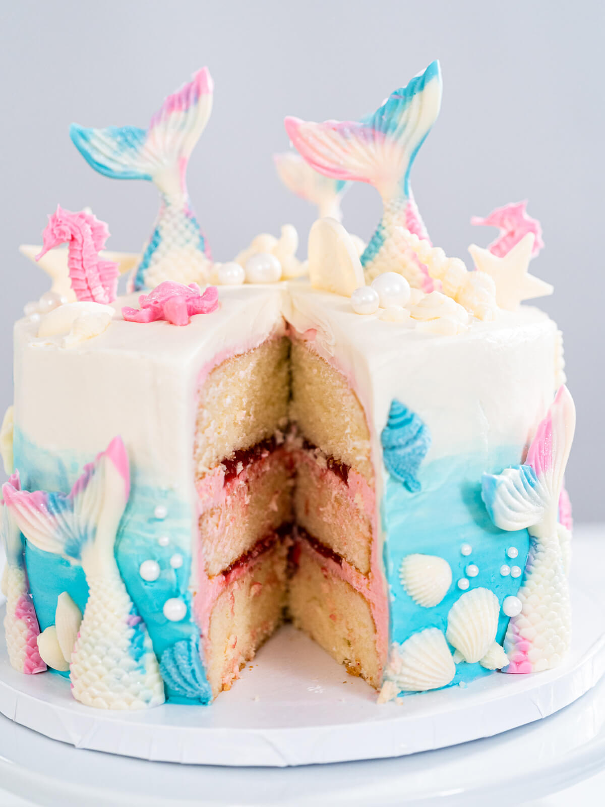 Meerjungfrauen-Torte mit herausgeschnittenem Stück, drei Vanilleschichten mit rosa Buttercreme und roter Konfitüre, außen blau-weißer Farbverlauf mit Schokoladenmuscheln
