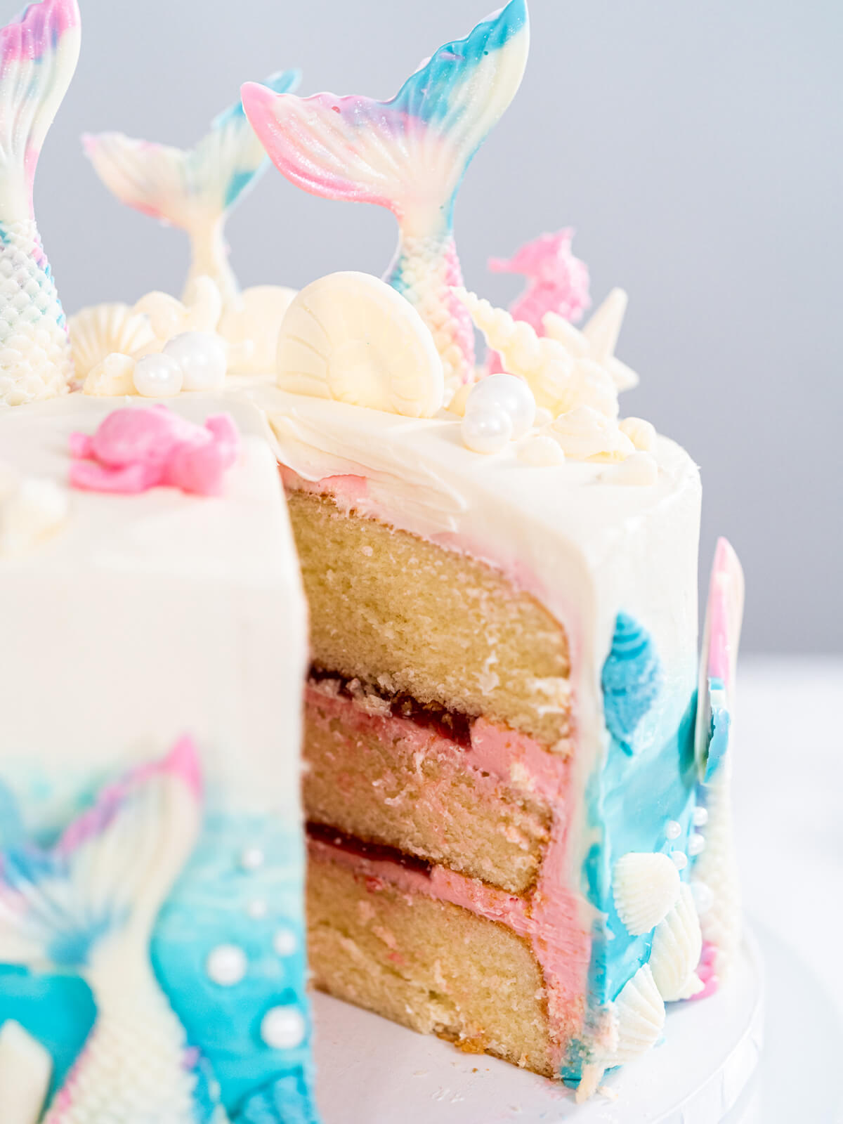 Angeschnittene Meerjungfrauen-Torte in Nahaufnahme, drei Vanilleschichten mit rosa Buttercreme und dunkelroter Konfitüre, oben Schokoladen-Flossen und weiße Zuckerperlen