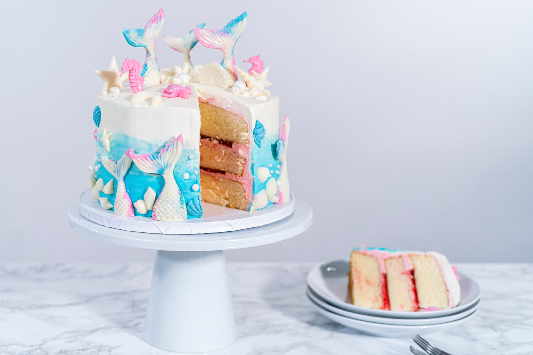 Angeschnittene Meerjungfrauen-Torte auf weißem Tortenständer, daneben ein Stück auf einem Tellerstapel mit sichtbaren Vanilleschichten und roter Konfitüre