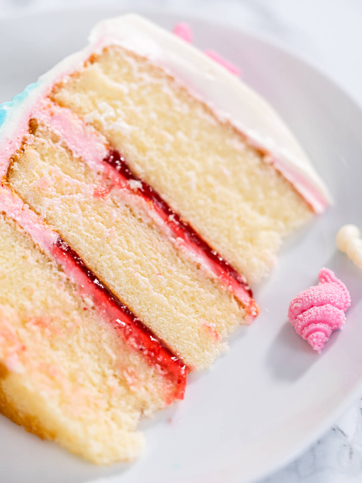 Angeschnittenes Stück Meerjungfrauen-Torte auf weißem Teller, drei helle Vanilleschichten mit rosa Buttercreme und roter Konfitüre, außen weißer Überzug