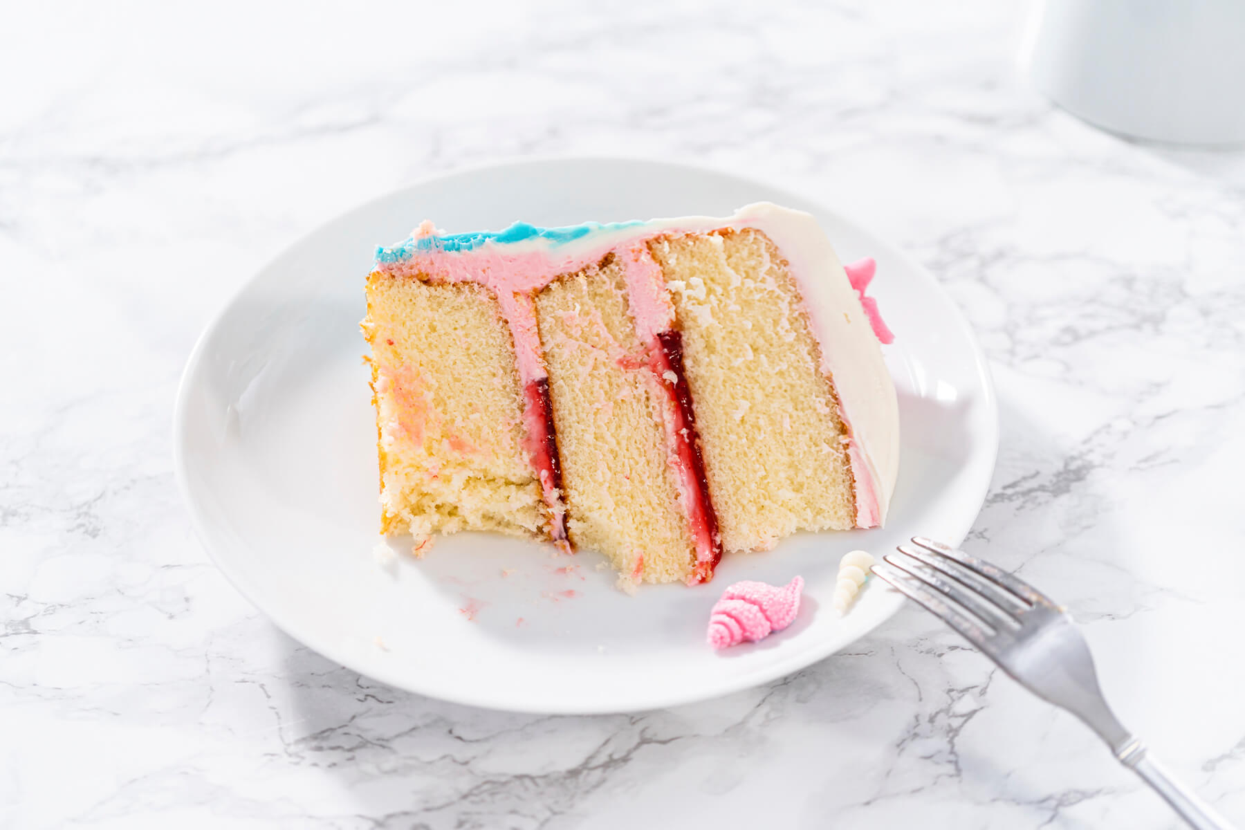 Ein Stück Meerjungfrauen-Torte auf weißem Teller mit Gabel, drei Vanilleschichten mit rosa Creme und roter Konfitüre, daneben eine rosa Schokoladenmuschel