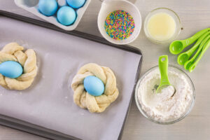 Aufsicht auf ein rundes Kränzchen aus zwei verdrehten Teigsträngen mit blau gefärbtem Ei tief in der Mitte, daneben ein Schälchen bunte Zuckerstäbchen, ein Schälchen zerlassene Butter und eine Schüssel Mehl