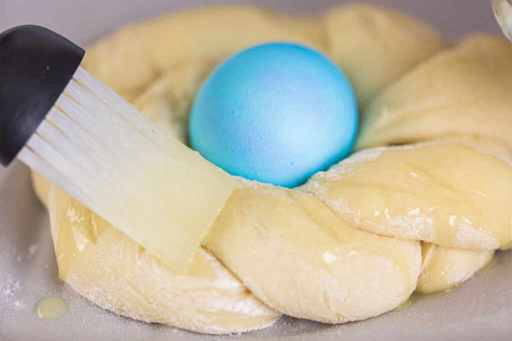Silikonpinsel streicht hell gelbe, zerlassene Butter auf die verdrehten Teigstränge eines Mini-Osterbrots, in der Mitte das kräftig blau gefärbte Ei, auf dem Blech ein Butter-Tropfen