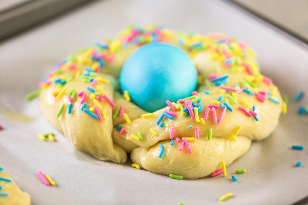 Mit Ei bestrichenes und dicht mit rosa, blauen, gelben und grünen Zuckerstäbchen bestreutes Hefekränzchen mit blauem Ei auf Backpapier, kurz vor dem Backen