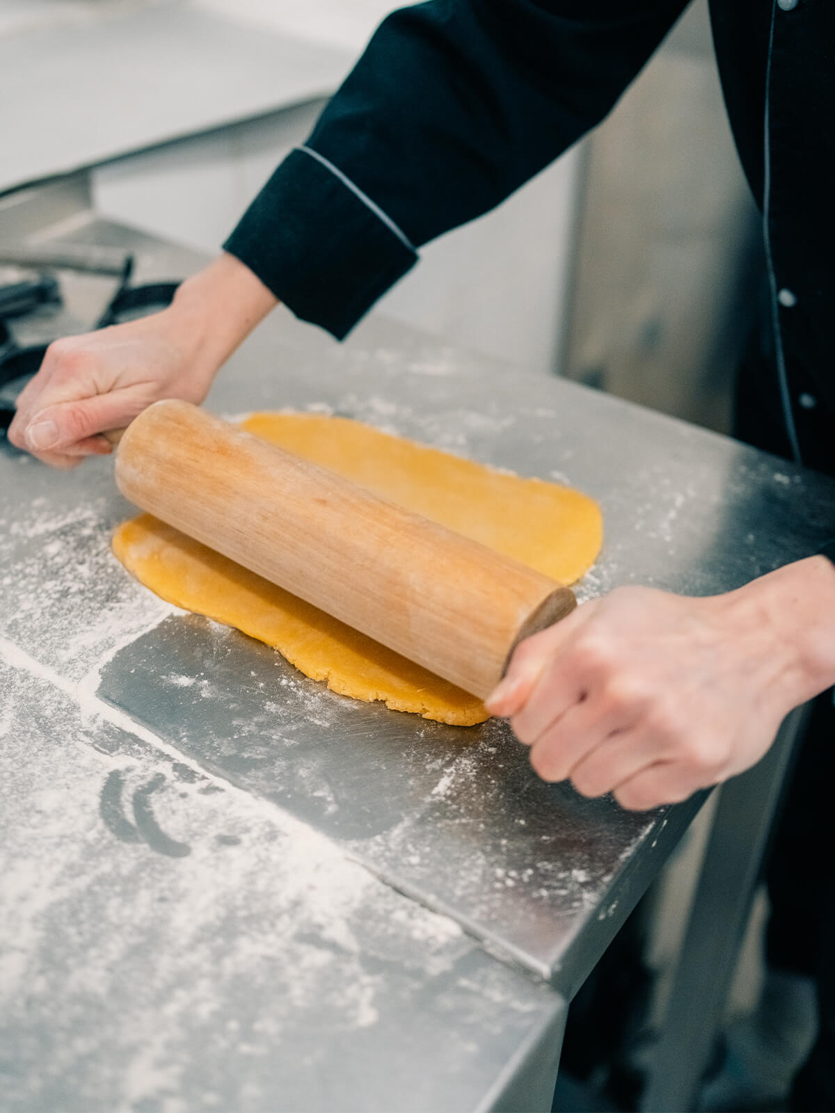 Hände rollen eine gelbe Mürbeteigplatte mit einem Holzrollholz auf einer bemehlten Edelstahlplatte aus