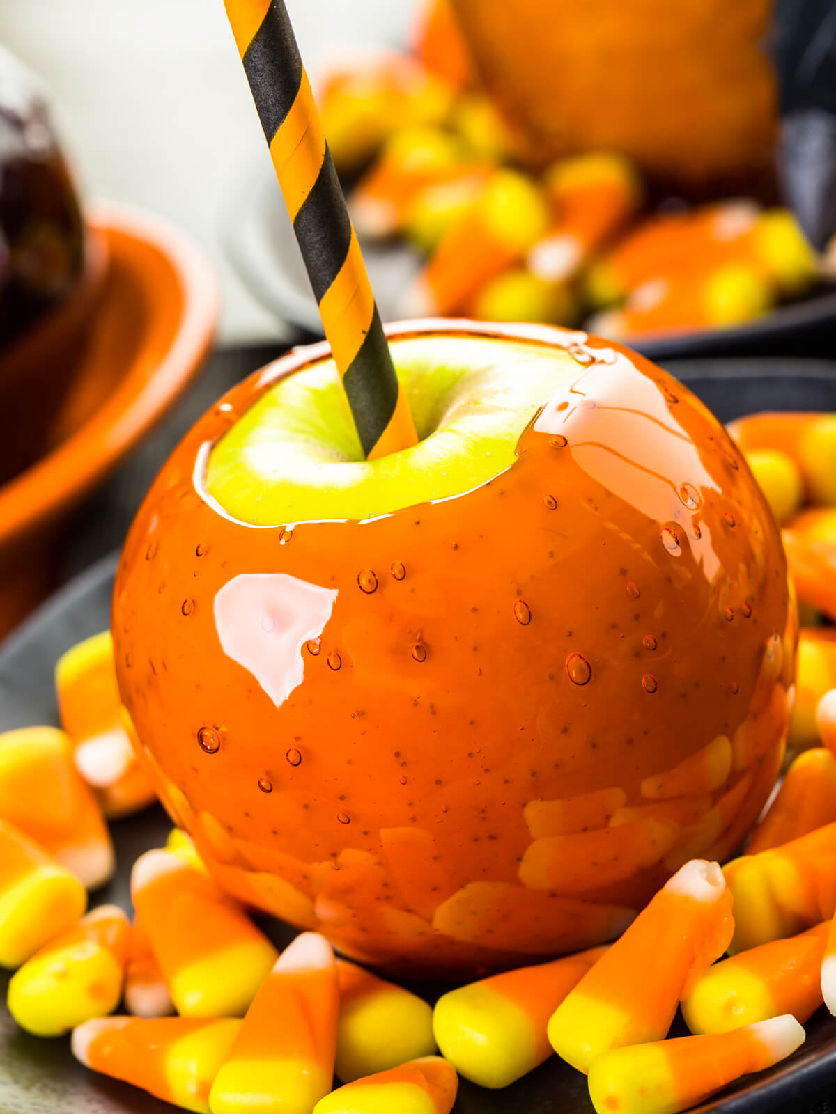 Oranger Liebesapfel im Hochformat aus kurzer Distanz, ringsum liegen dreifarbige Bonbons; die Glasur läuft über die Schulter bis an den Rand der Stielmulde und bildet dort einen dicken Wulst, obenauf bleibt rund um den orange-schwarz gestreiften Halm eine blanke grüne Fläche frei, in der Schicht stehen große und kleine Luftbläschen, hinten unscharf ein heller Kürbis und ein weiterer Apfel