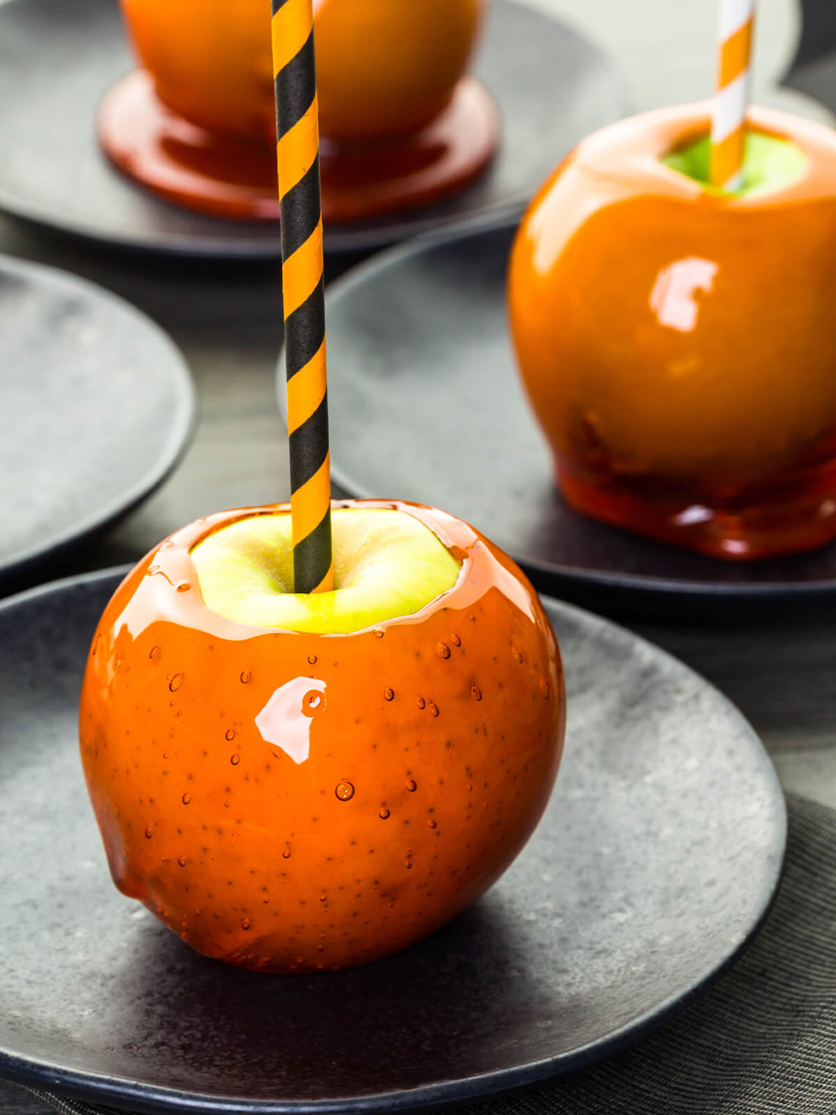 Mehrere orange Liebesäpfel auf jeweils eigenen matten schwarzen Tellern, gestaffelt in die Tiefe; der vordere Apfel zeigt eine kürbisorange, spiegelnde Glasur mit Luftbläschen und einem hellen Reflex, obenauf rund um den orange-schwarz gestreiften Papierstrohhalm eine blanke grüne Fläche von etwa halber Apfelbreite, bei den hinteren Äpfeln läuft die Glasur am Fuß zu einer breiten glänzenden Scheibe aus, der rechte trägt einen orange-weiß gestreiften Halm