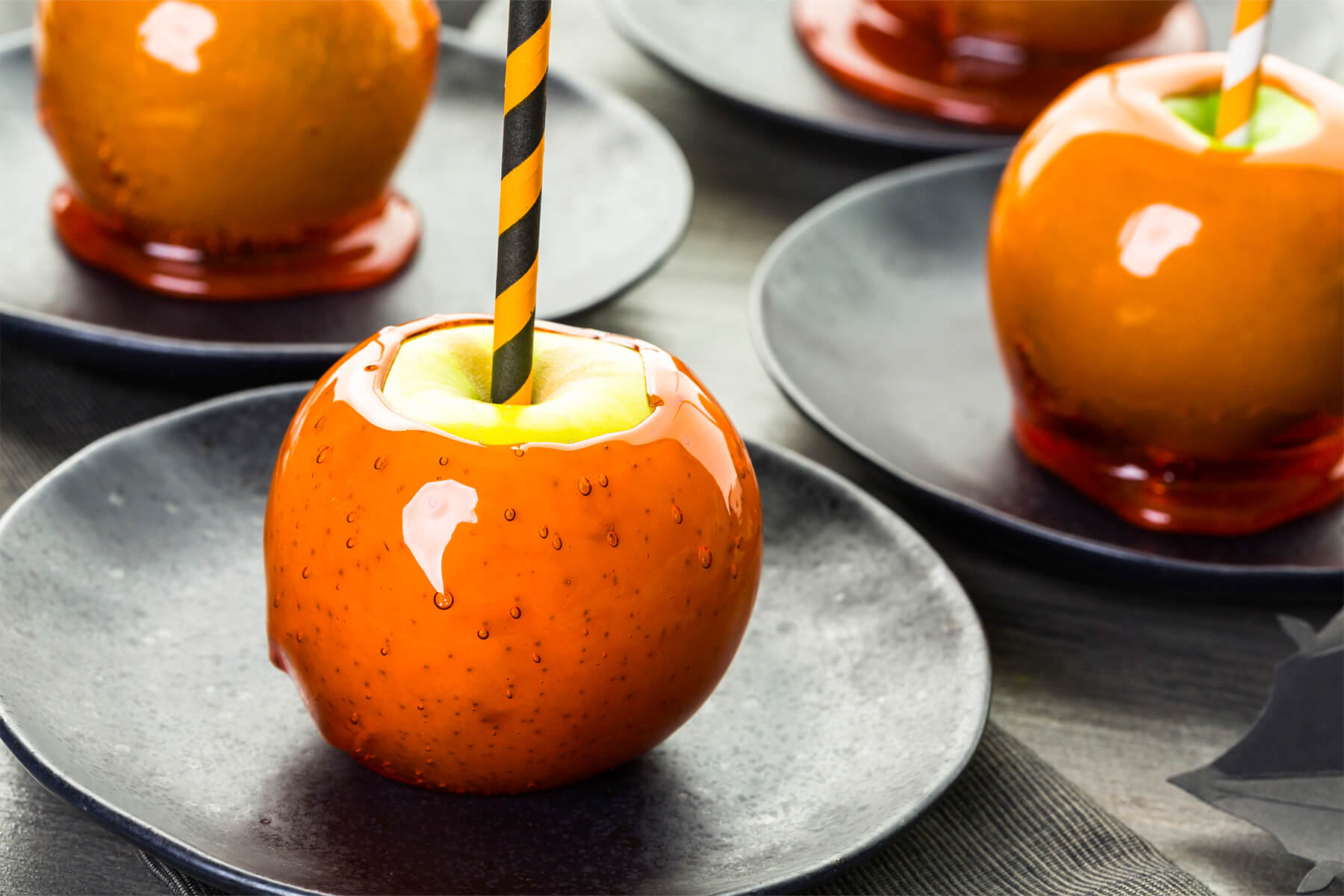 Blick über mehrere matte schwarze Teller mit orangen Liebesäpfeln, vorn ein Apfel in voller Schärfe: die Glasur läuft am Fuß zu einer flachen glänzenden Scheibe aus, in der Schicht stehen dicht an dicht Luftbläschen von Stecknadelkopf- bis Linsengröße, obenauf eine blanke grüne Fläche rund um den orange-schwarz gestreiften Halm, rechts hinten ein Apfel mit orange-weißem Halm