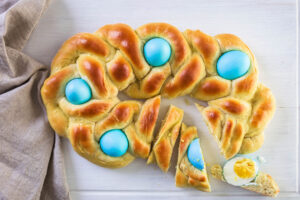 Geflochtenes italienisches Osterbrot als ovaler Ring von oben mit fünf blau gefärbten Eiern, rechts in Scheiben geschnitten, daneben ein halbiertes hart gekochtes Ei auf einem Stück Brot