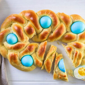 Geflochtenes italienisches Osterbrot als ovaler Ring von oben mit fünf blau gefärbten Eiern, rechts in Scheiben geschnitten, daneben ein halbiertes hart gekochtes Ei auf einem Stück Brot