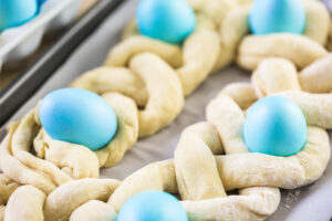 Nahaufnahme des aufgegangenen Flechtrings mit blau gefärbten Eiern, die bis etwa zur Hälfte in den Teiglücken sitzen, noch ungebacken auf dem Blech