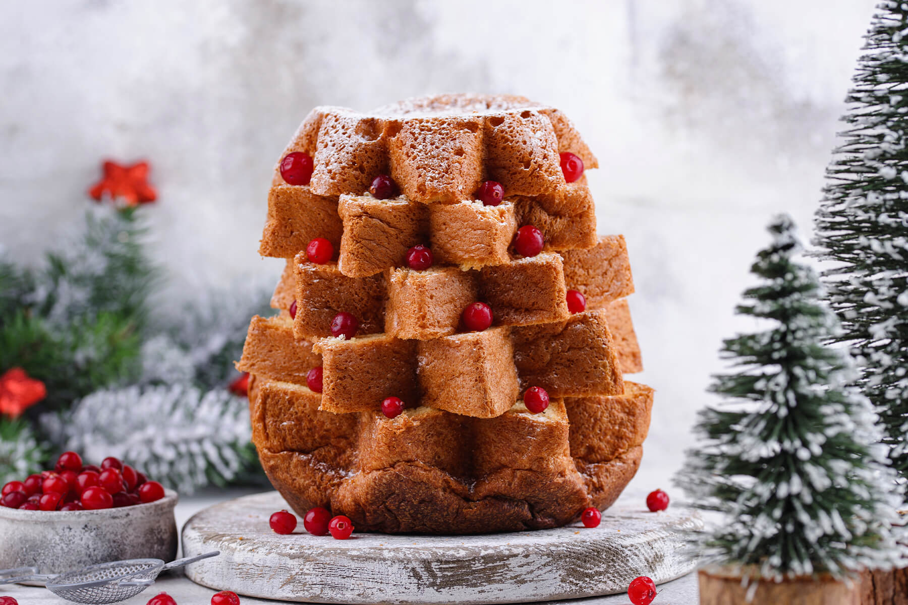 Pandoro-Baum aus versetzten Sternscheiben ohne Creme, rote Beeren liegen in den Zackenlücken, ringsum verschneite Deko-Tannen