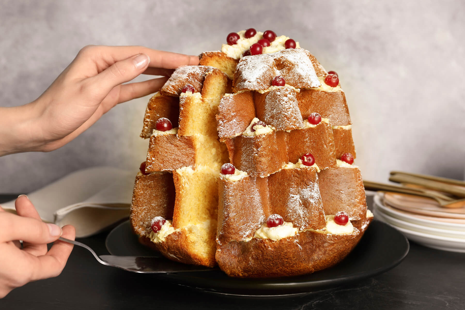 Zwei Hände heben mit einem Tortenheber einen senkrechten Keil aus einem Pandoro-Baum, die Schnittfläche zeigt helle Krume und Creme