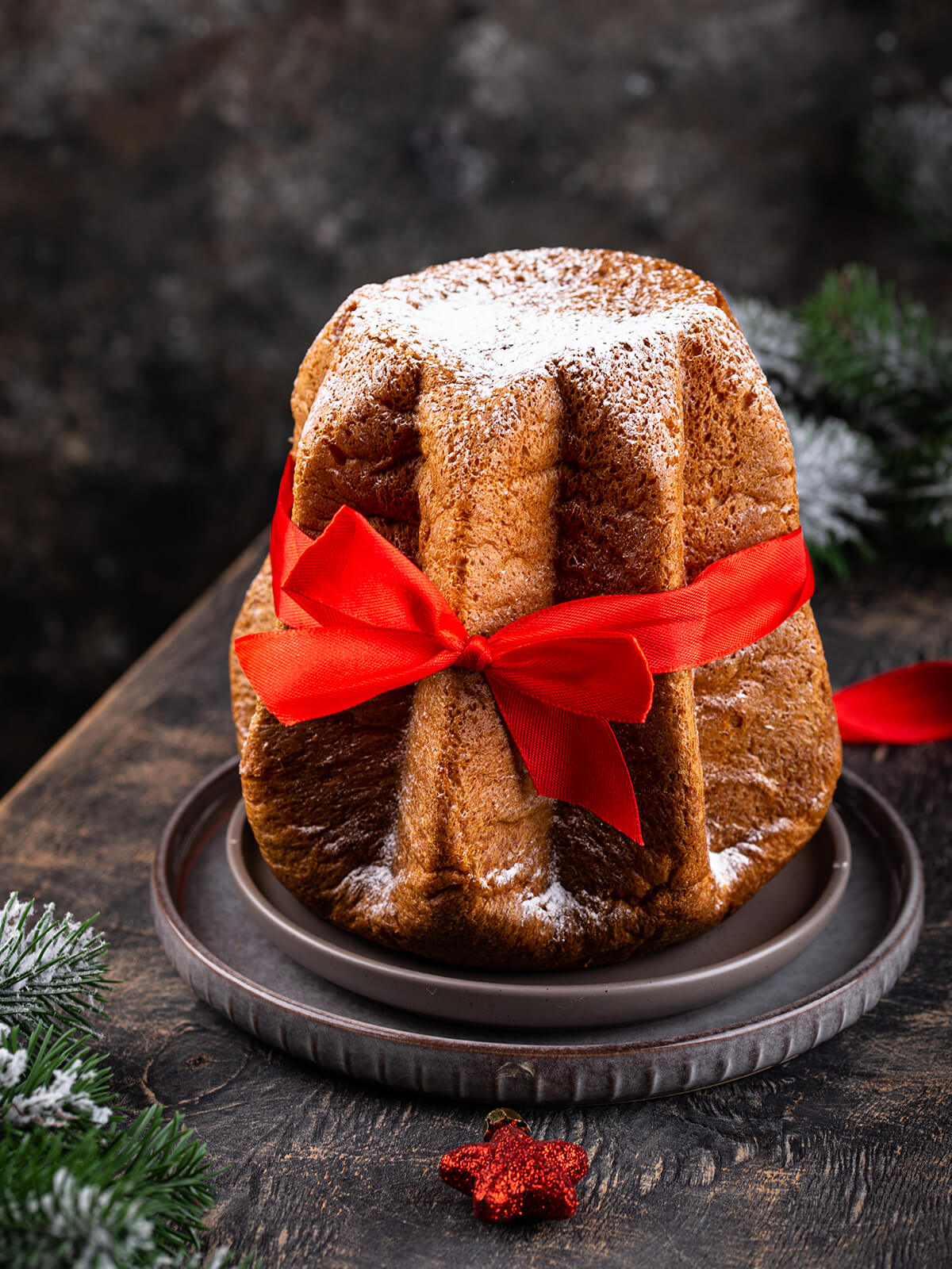 Ganzer Pandoro mit Puderzucker bestäubt und mit einer roten Satinschleife umbunden, daneben verschneite Tannenzweige