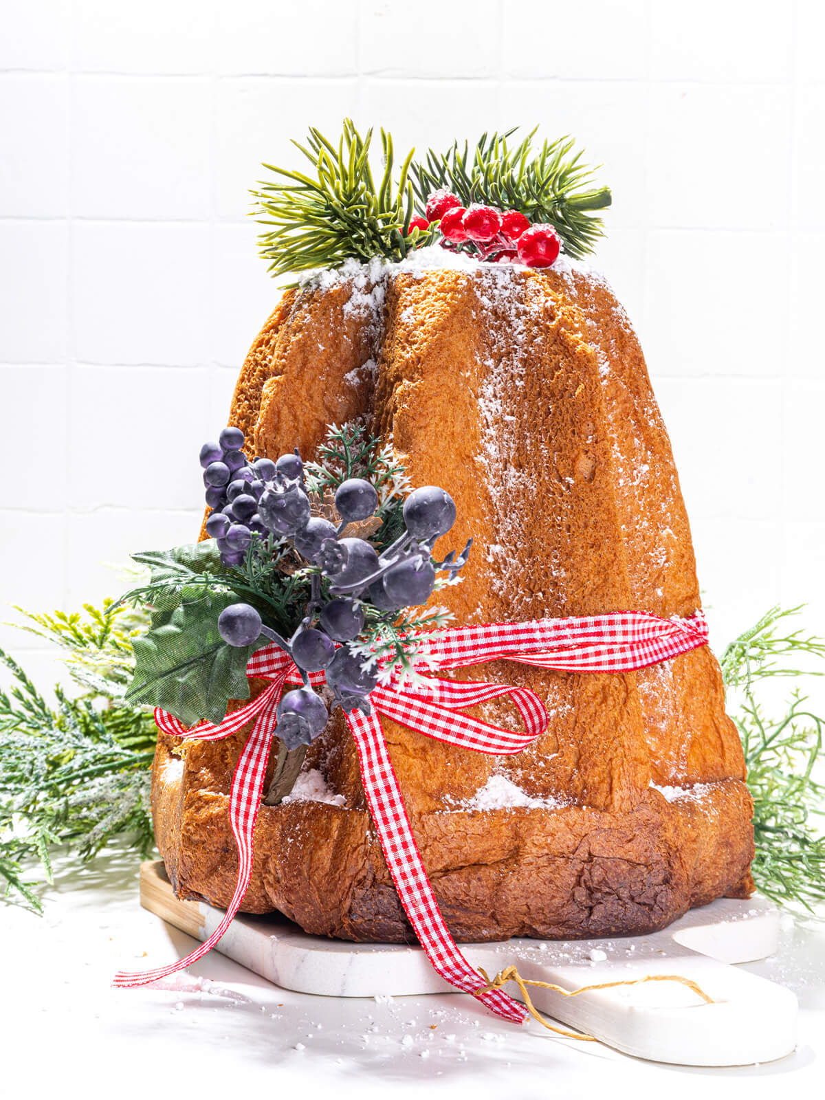 Pandoro mit Karoband, künstlichen Beerenzweigen auf Draht und synthetischer Deko-Tanne vor weißer Wand