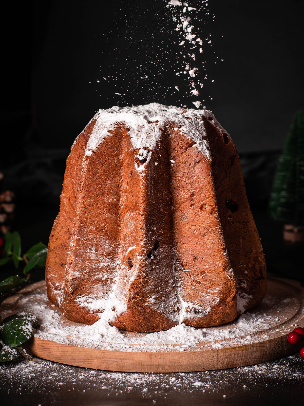 Puderzucker rieselt aus der Höhe auf einen goldbraunen Pandoro auf einem runden Holzbrett vor dunklem Hintergrund