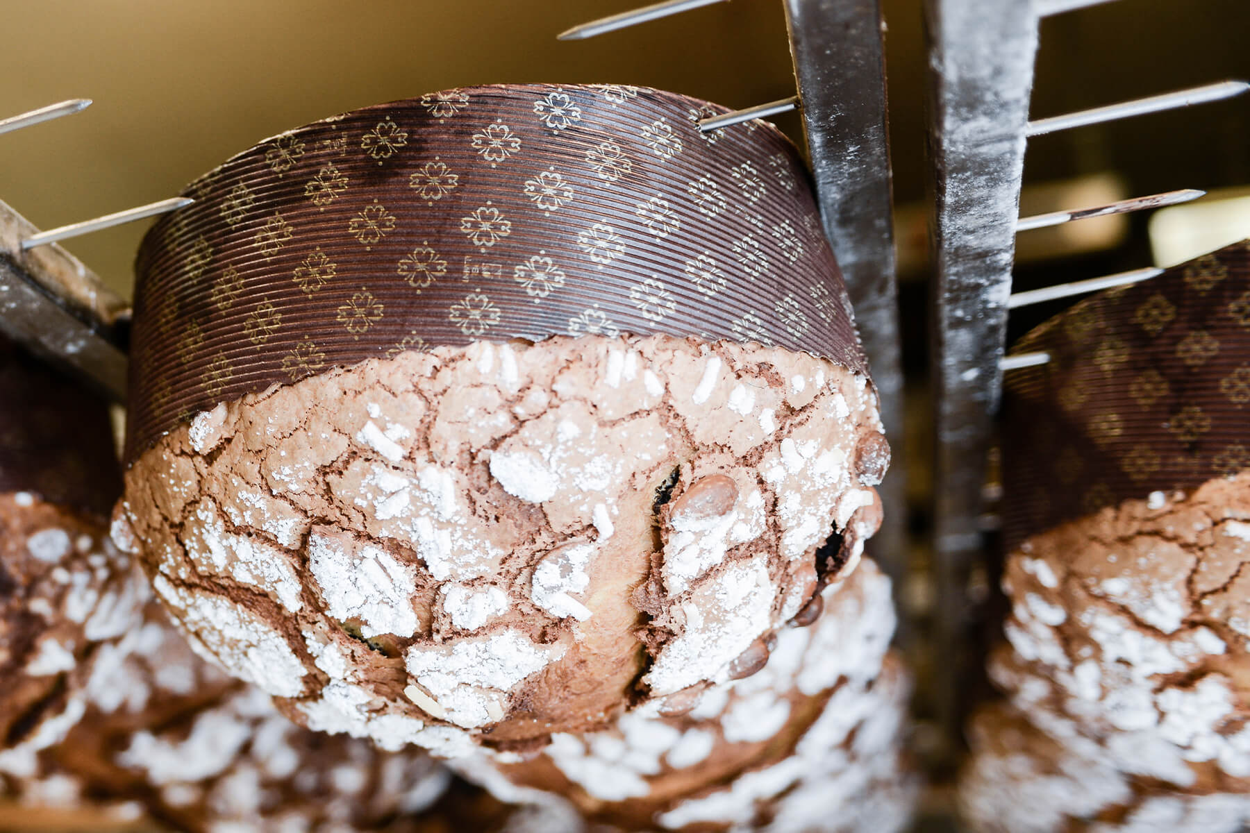 Nahaufnahme eines kopfüber hängenden Panettone, zwei Metallspieße stecken quer durch die Papierform, die Glasur hängt nach unten