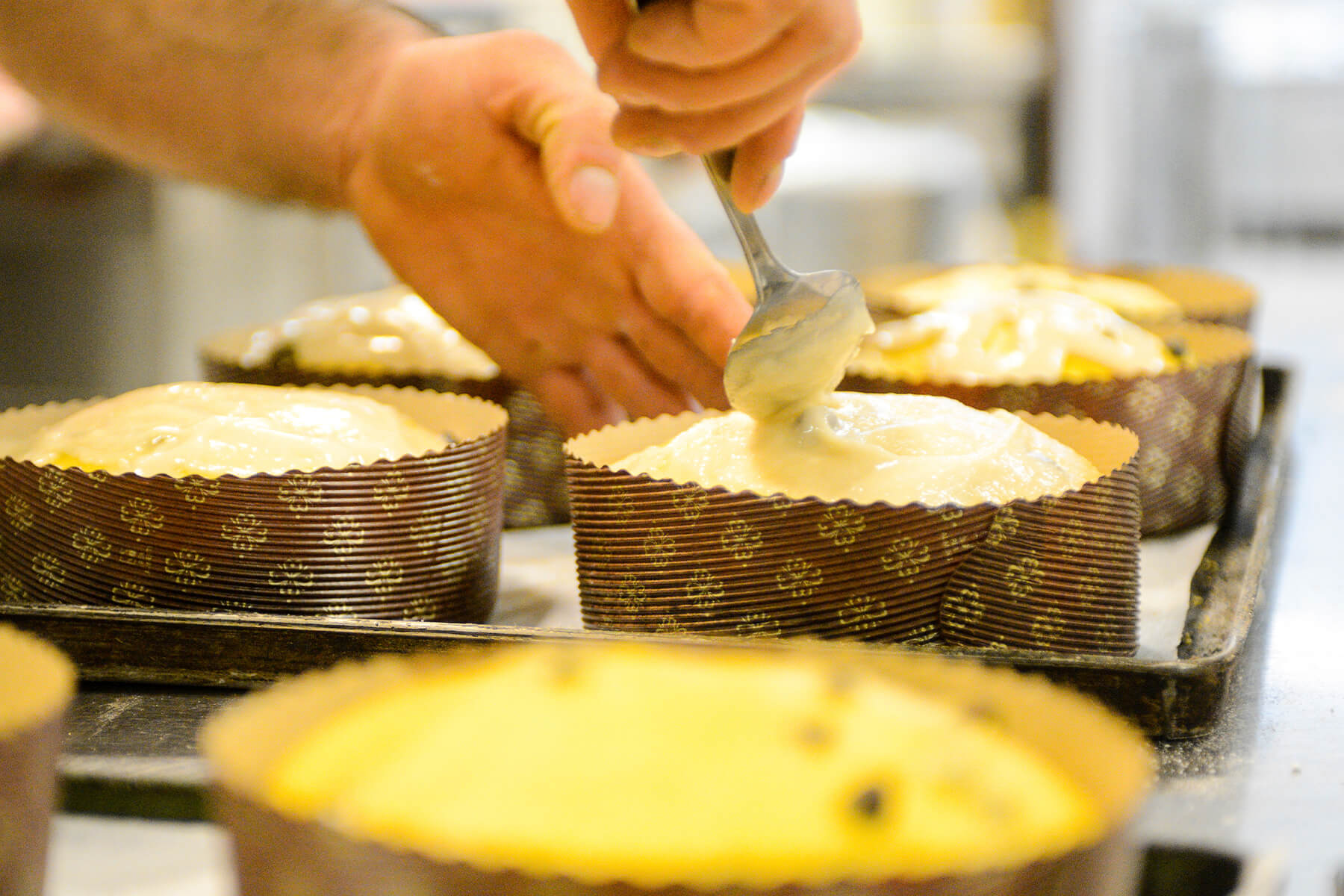 Eine Hand streicht mit einem Löffel helle Mandelglasur auf den rohen, aufgegangenen Panettone-Teig in der Papierform