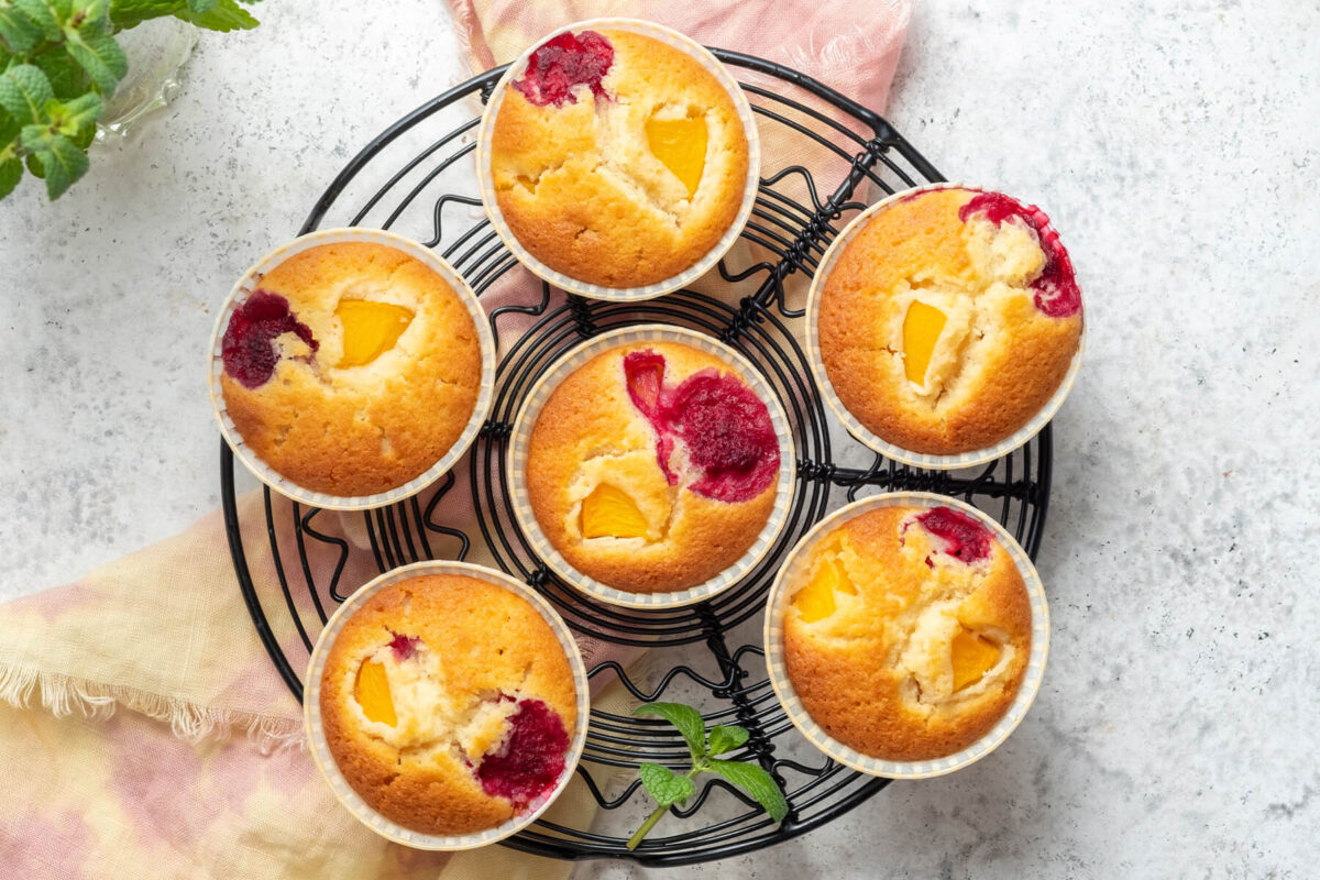 Sechs Pfirsich-Himbeer-Muffins von oben auf einem schwarzen Kuchengitter, jeder mit einem gelben Pfirsichstück und einer roten Himbeere an der Oberfläche
