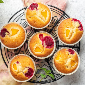 Sechs Pfirsich-Himbeer-Muffins von oben auf einem schwarzen Kuchengitter, jeder mit einem gelben Pfirsichstück und einer roten Himbeere an der Oberfläche