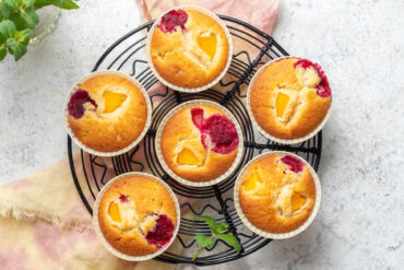 Sechs Pfirsich-Himbeer-Muffins von oben auf einem schwarzen Kuchengitter, jeder mit einem gelben Pfirsichstück und einer roten Himbeere an der Oberfläche