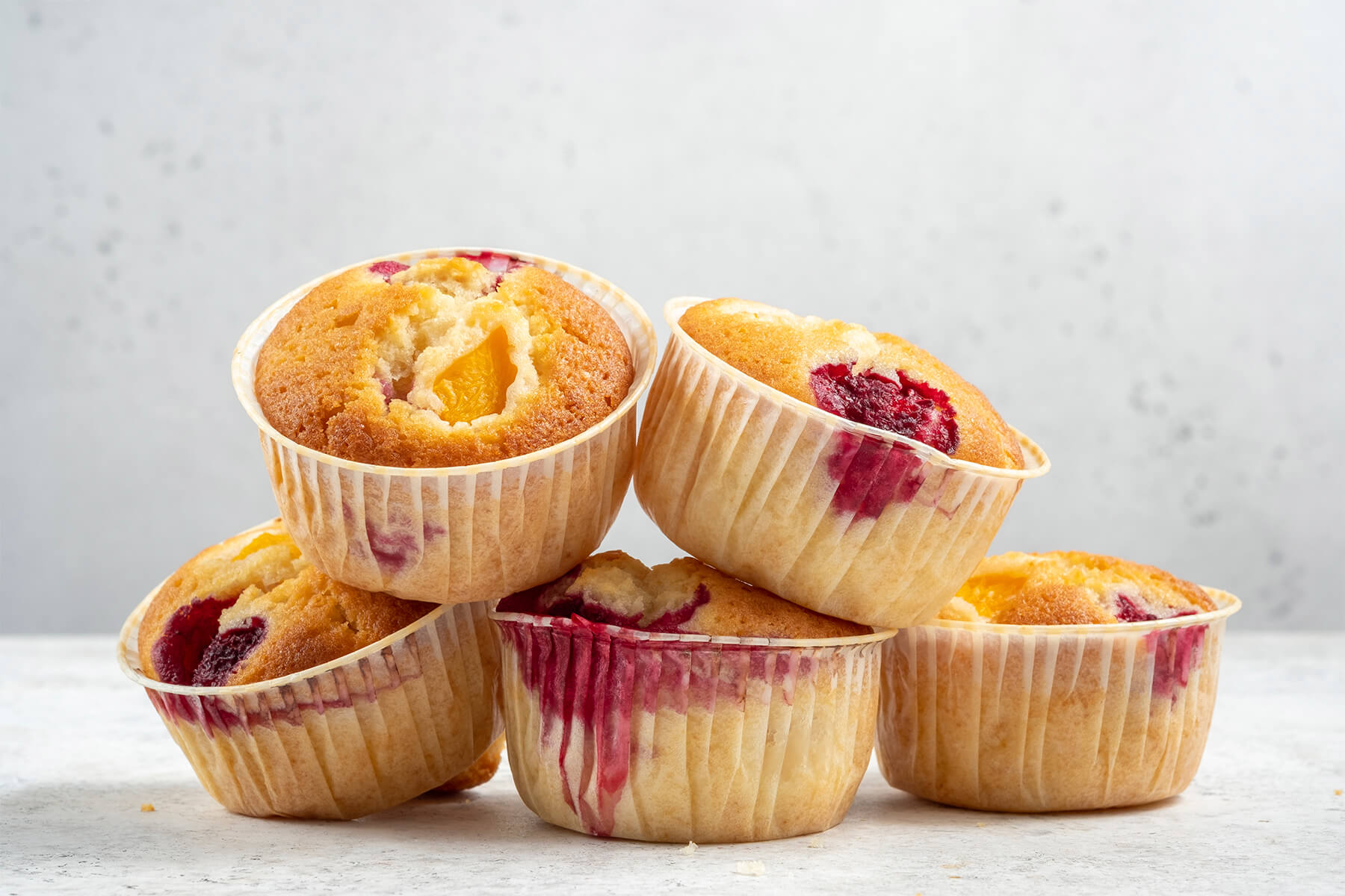 Fünf Muffins übereinandergestapelt, an den Oberseiten sind gelbe Pfirsichstücke und rote Himbeeren eingebacken