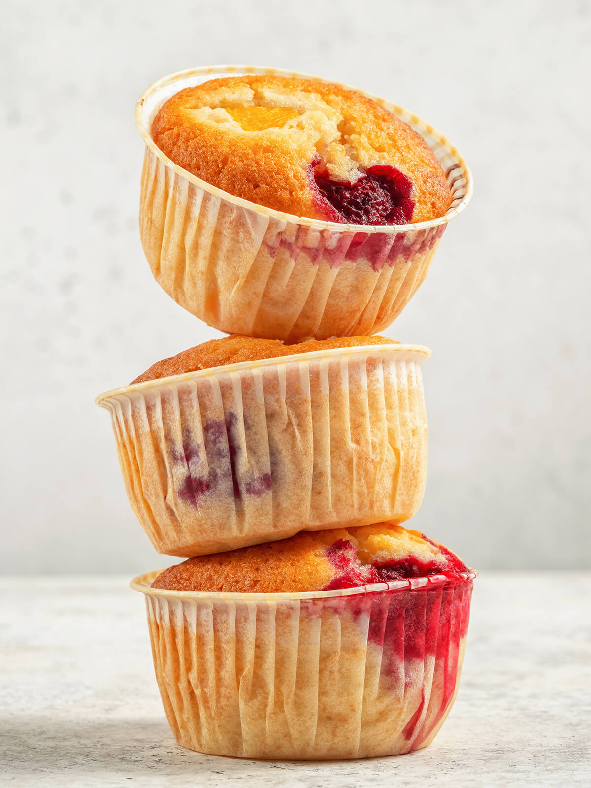 Drei gestapelte Muffins in durchscheinenden Papierförmchen, durch das Papier sind rote Himbeerspuren über die ganze Höhe zu sehen