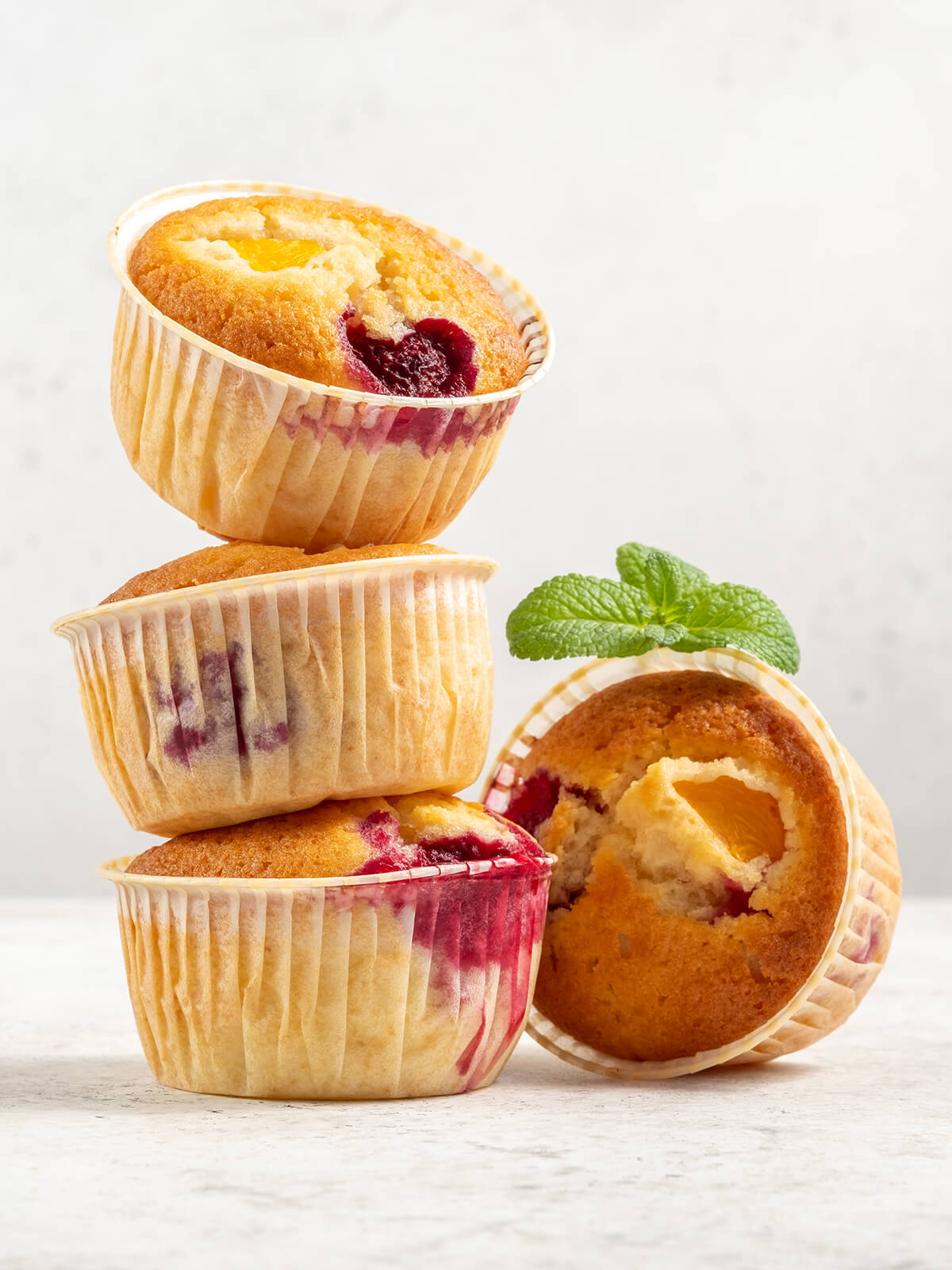 Gestapelte Muffins mit fruchtigen Spuren im Papier, davor ein angelehnter Muffin mit sichtbarem Pfirsichstück und ein Minzzweig