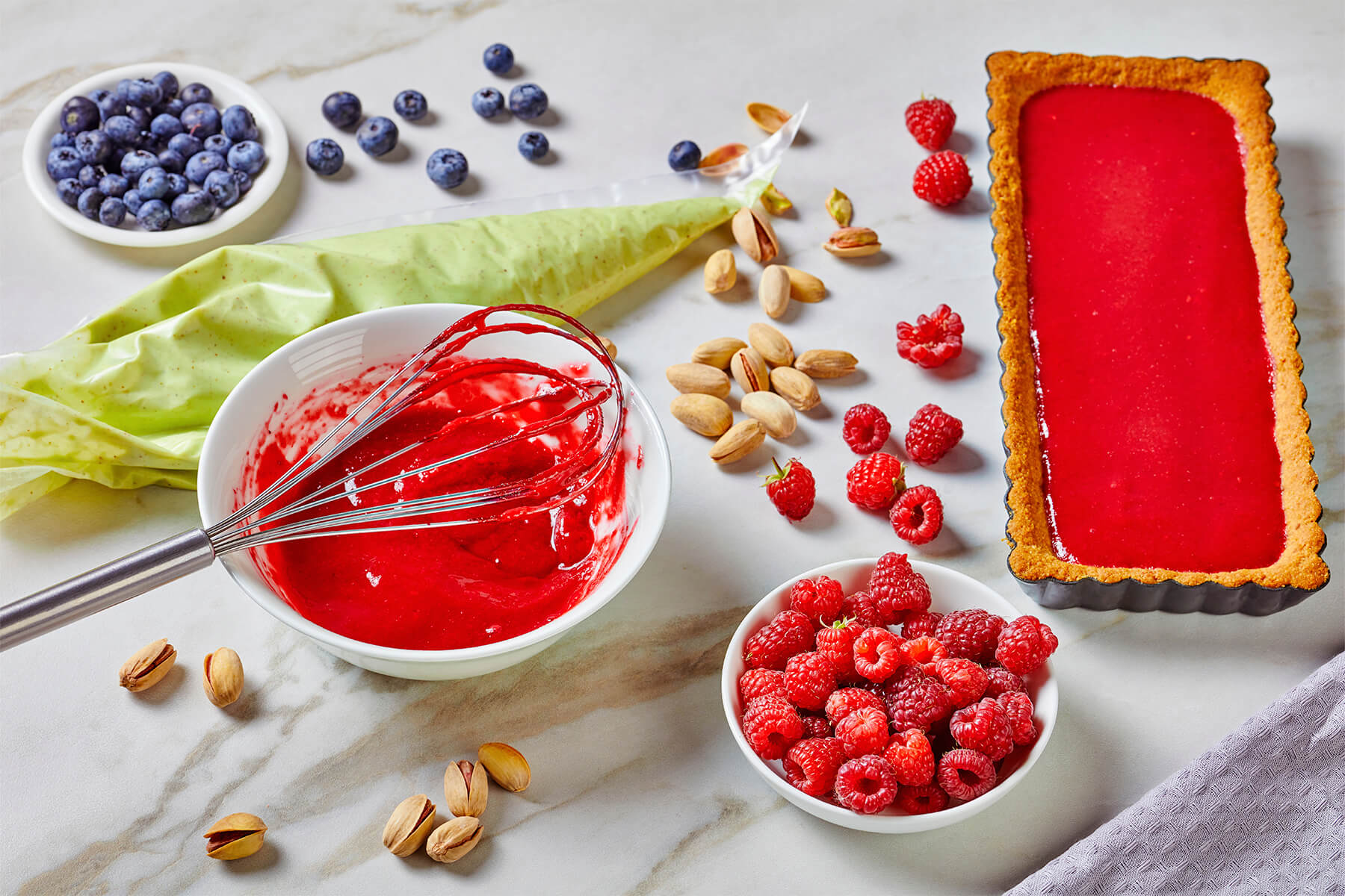 Ein gefüllter Spritzbeutel mit heller grüner Creme neben einer Schüssel Fruchtpüree, Beeren und Nüssen auf einer Marmorplatte