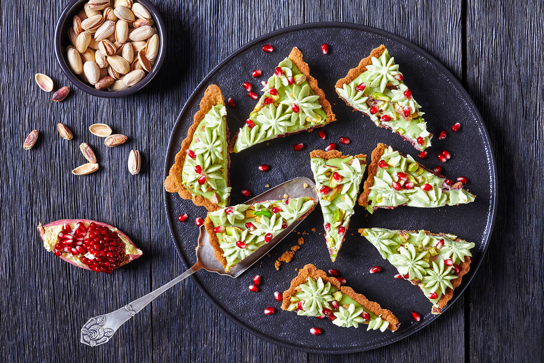 In acht Stücke geschnittene runde Tarte auf einem schwarzen Teller, an den Schnittkanten ist unter der grünen Creme eine dünne rote Geleeschicht zu sehen