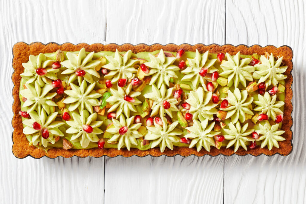 Rechteckige Pistazien-Ganache-Tarte von oben, dicht mit hellgrünen Sahnerosetten, Granatapfelkernen und gehackten Pistazien belegt