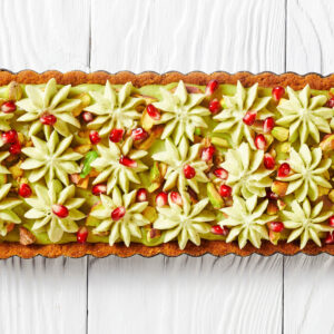 Rechteckige Pistazien-Ganache-Tarte von oben, dicht mit hellgrünen Sahnerosetten, Granatapfelkernen und gehackten Pistazien belegt