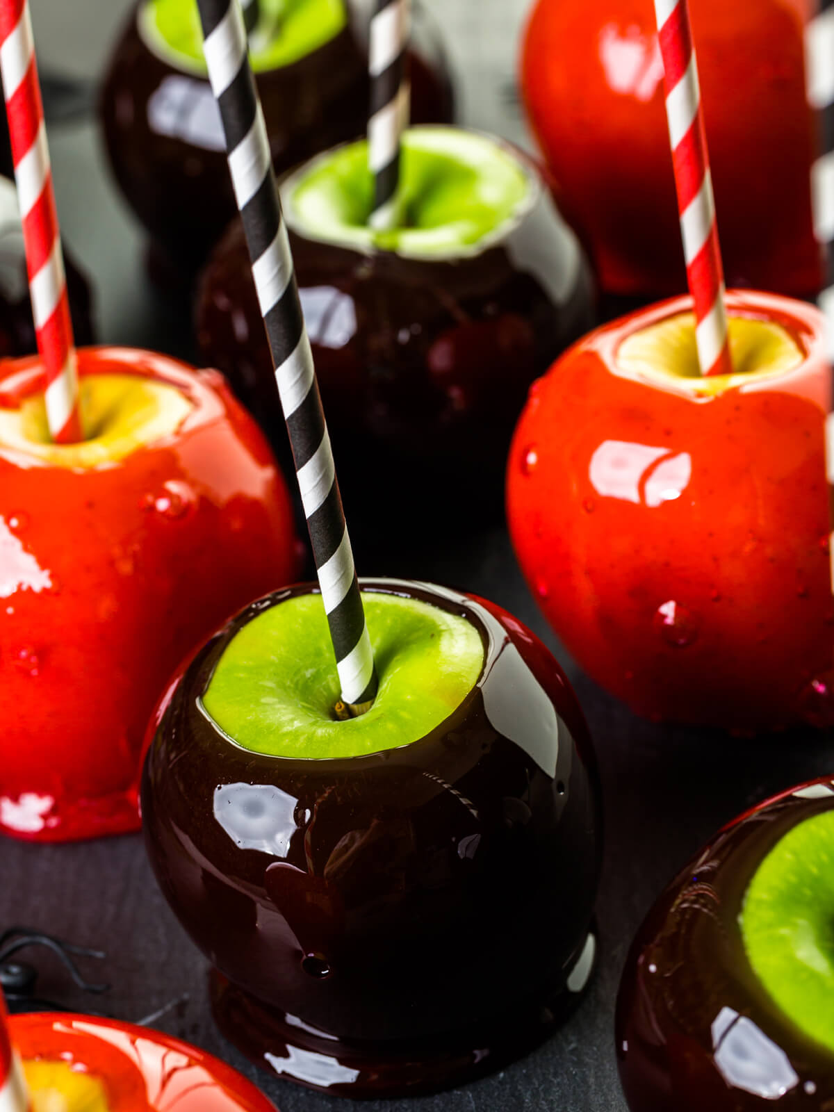 Mehrere Liebesäpfel dicht gedrängt auf dunklem Untergrund, die Glasur spiegelt das Licht in scharf abgegrenzten weißen Reflexen; links und rechts leuchtend rote Äpfel an rot-weiß gestreiften Papierstrohhalmen, in deren Stielmulde ein Ring gelber Apfelschale frei liegt und in deren roter Schicht viele winzige Luftbläschen stehen, vorn ein fast schwarz durchscheinender Apfel an einem schwarz-weißen Halm über grüner Schale
