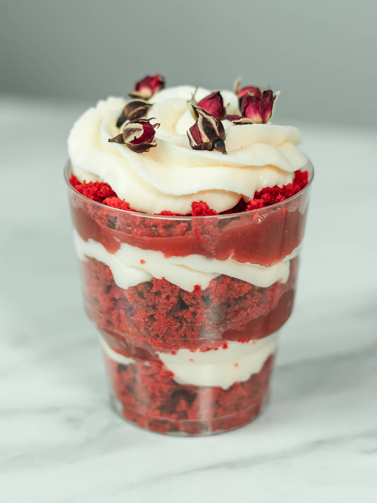 Rotes Schichtdessert im Glas mit weißer Cremekrone und getrockneten Rosenknospen
